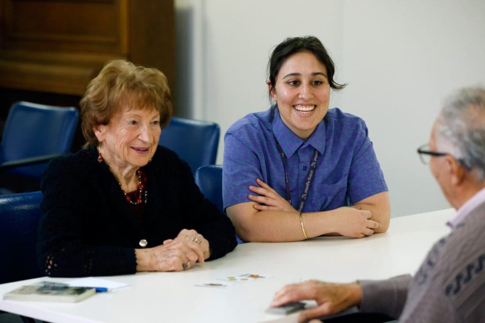 Three people sit together at a table during a card game or social activity in a bright communal room. A staff member wearing a purple uniform sits alongside two older adults, with playing cards visible on the table as they engage in conversation and the activity together.