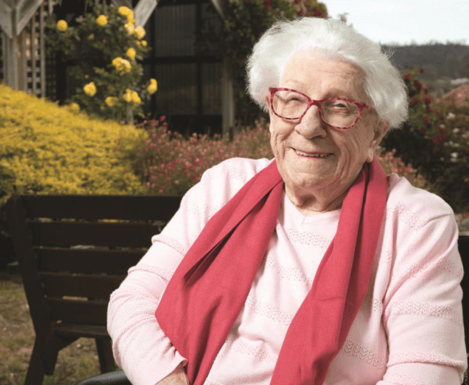 A person wearing glasses and a light pink jumper with a matching scarf sits outdoors in a garden setting. Colourful flowering shrubs and landscaped greenery surround a timber bench in the background, creating a peaceful and welcoming outdoor environment.