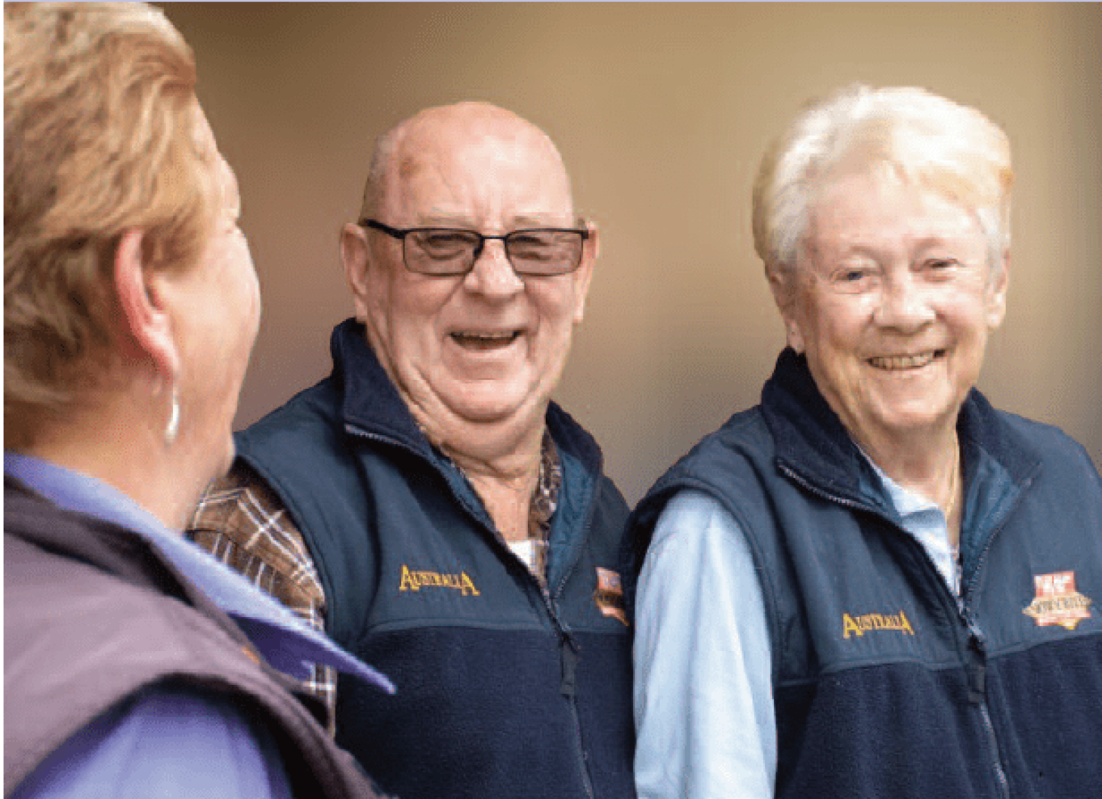 Two older adults wearing matching navy vests stand together and smile while speaking with a staff member in a purple uniform. The group is engaged in conversation in an indoor setting, creating a warm and welcoming sense of community and social connection.