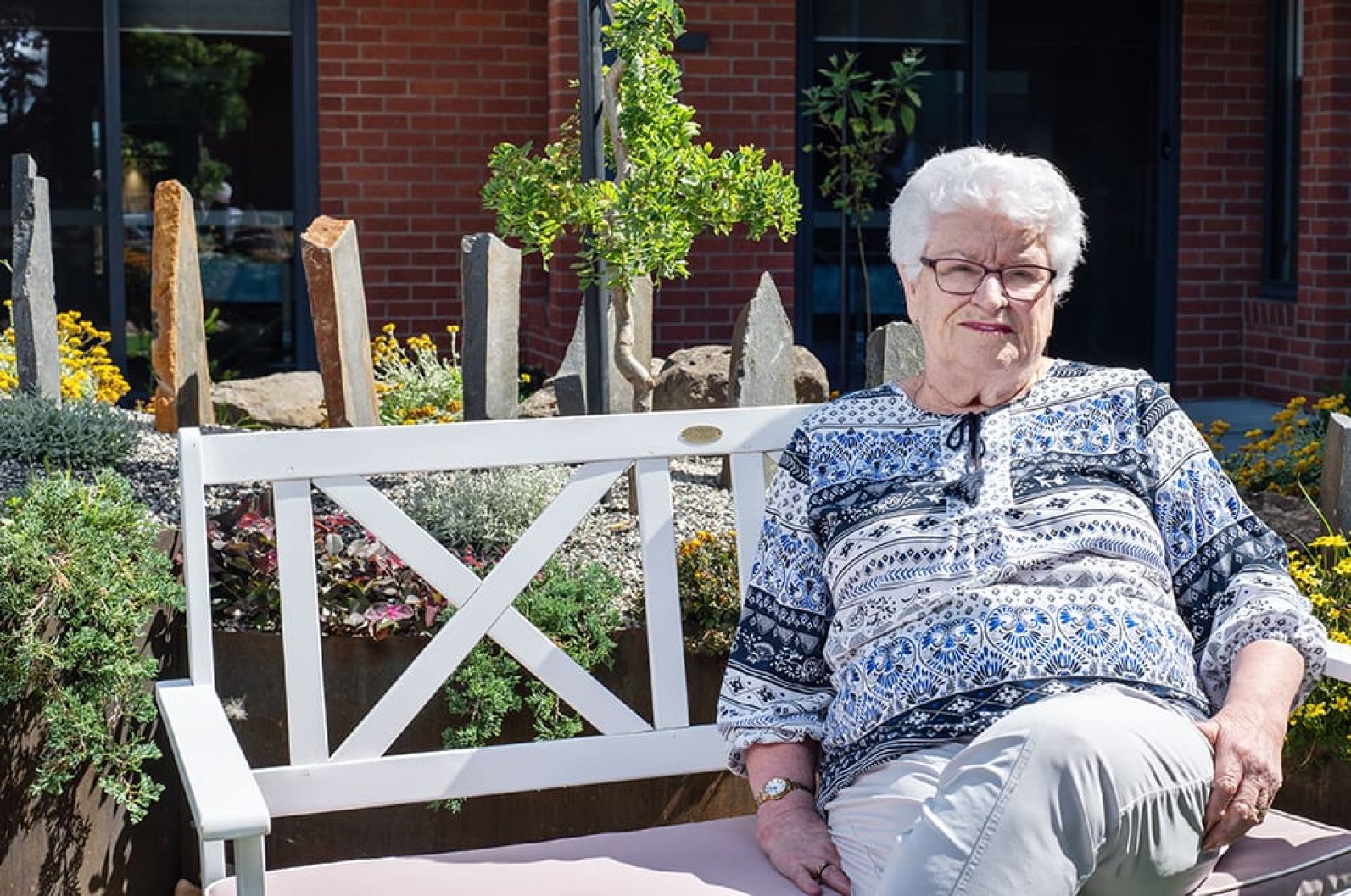 A person sits on a white outdoor bench in a landscaped courtyard garden. Flowering plants, shrubs and decorative stone features surround the seating area, while a red-brick building forms the background. The sunny outdoor setting provides a peaceful space for relaxation and enjoying the garden.