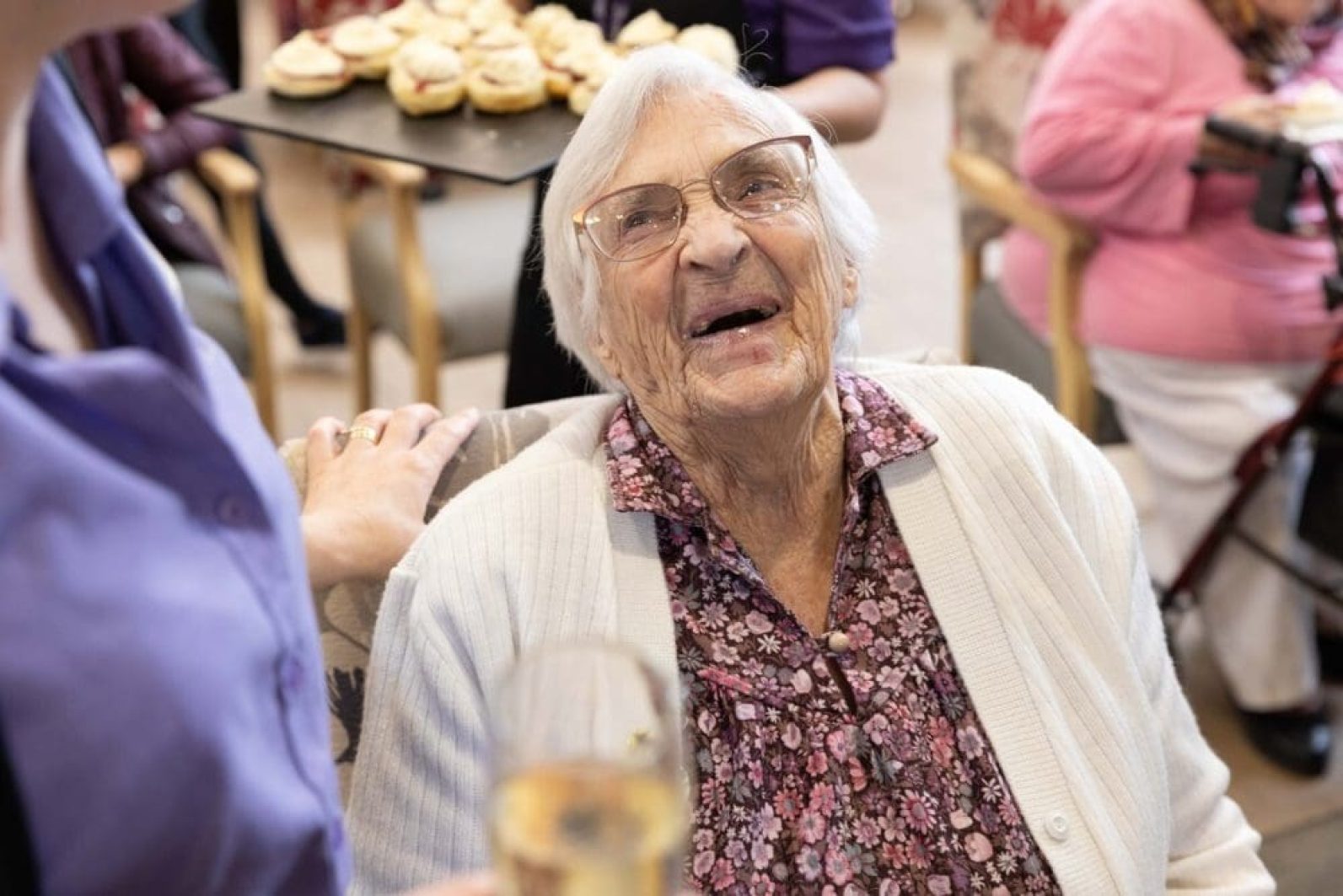 An older adult wearing glasses and a floral blouse sits in a chair during a social gathering, looking up while speaking with a staff member standing nearby. Other people are seated in the background, and a tray of small cakes is being served, creating a warm and welcoming community atmosphere.