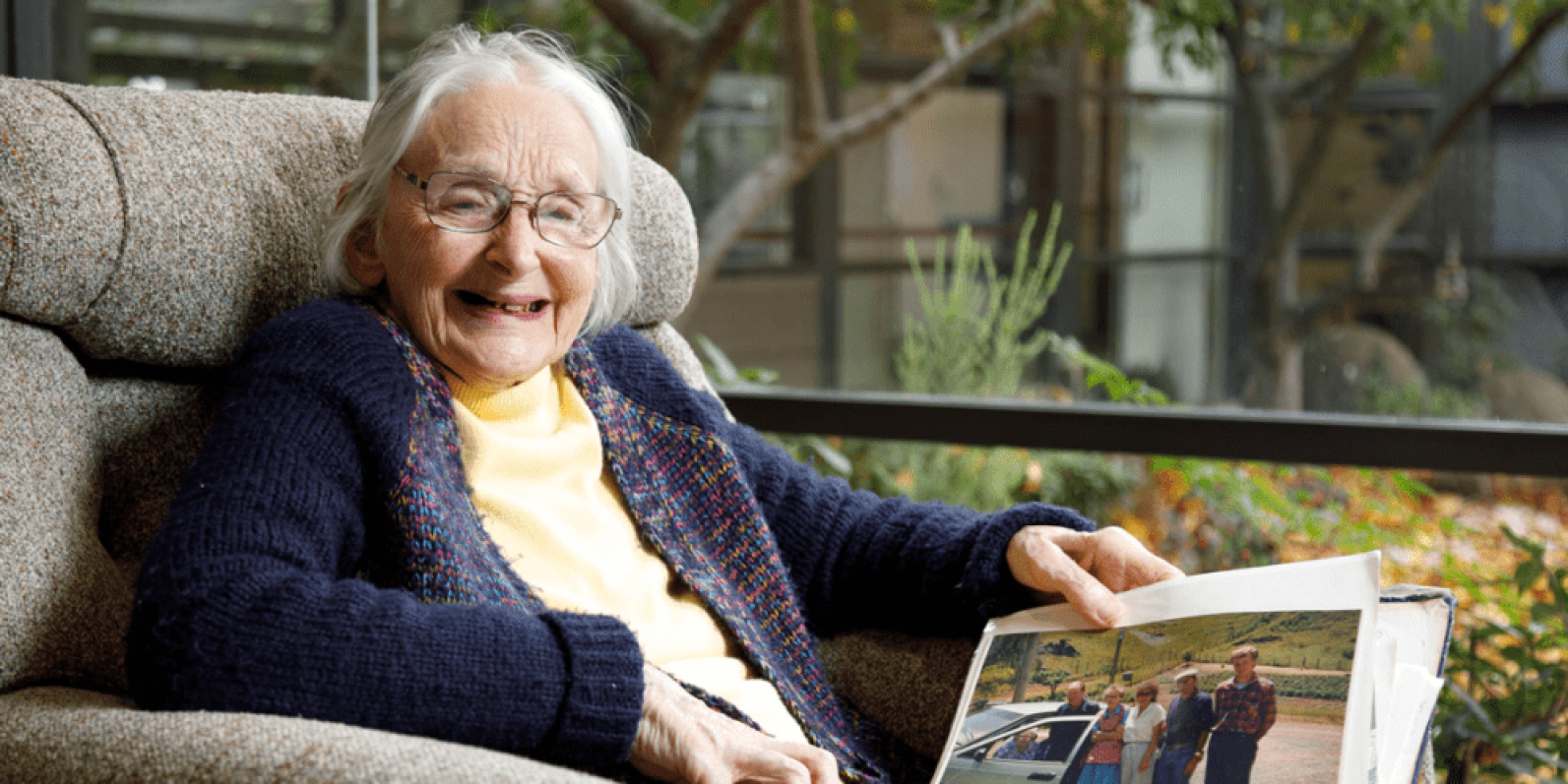 A person wearing glasses sits comfortably in a lounge chair beside a large window, looking through a photo album. Trees and garden greenery are visible outside, while soft furnishings and natural light create a warm and welcoming indoor setting.
