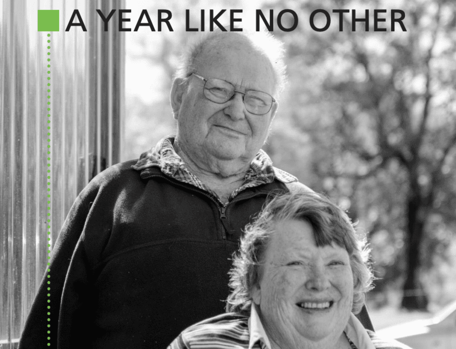 A black-and-white photograph shows two older adults posing outdoors, with one standing behind the other. The image includes the headline “A YEAR LIKE NO OTHER” across the top and a green graphic element along the left side. Trees and natural surroundings are softly blurred in the background.