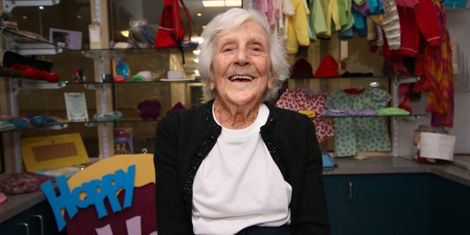 A person sits indoors in front of display shelves filled with colourful handmade clothing, accessories and craft items. The person is seated in a welcoming community craft or gift shop setting, with a sign reading “Happy” partially visible in the foreground and shelves of knitted and sewn items in the background.