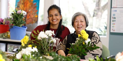 Two volunteers are pictured with white and yellow flowers in front of them.