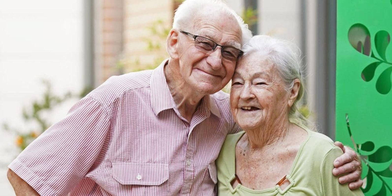 Two older adults stand close together outdoors, with one person’s arm around the other’s shoulder. The pair are positioned in front of a building entrance and greenery, creating a warm and welcoming scene that highlights companionship and connection.