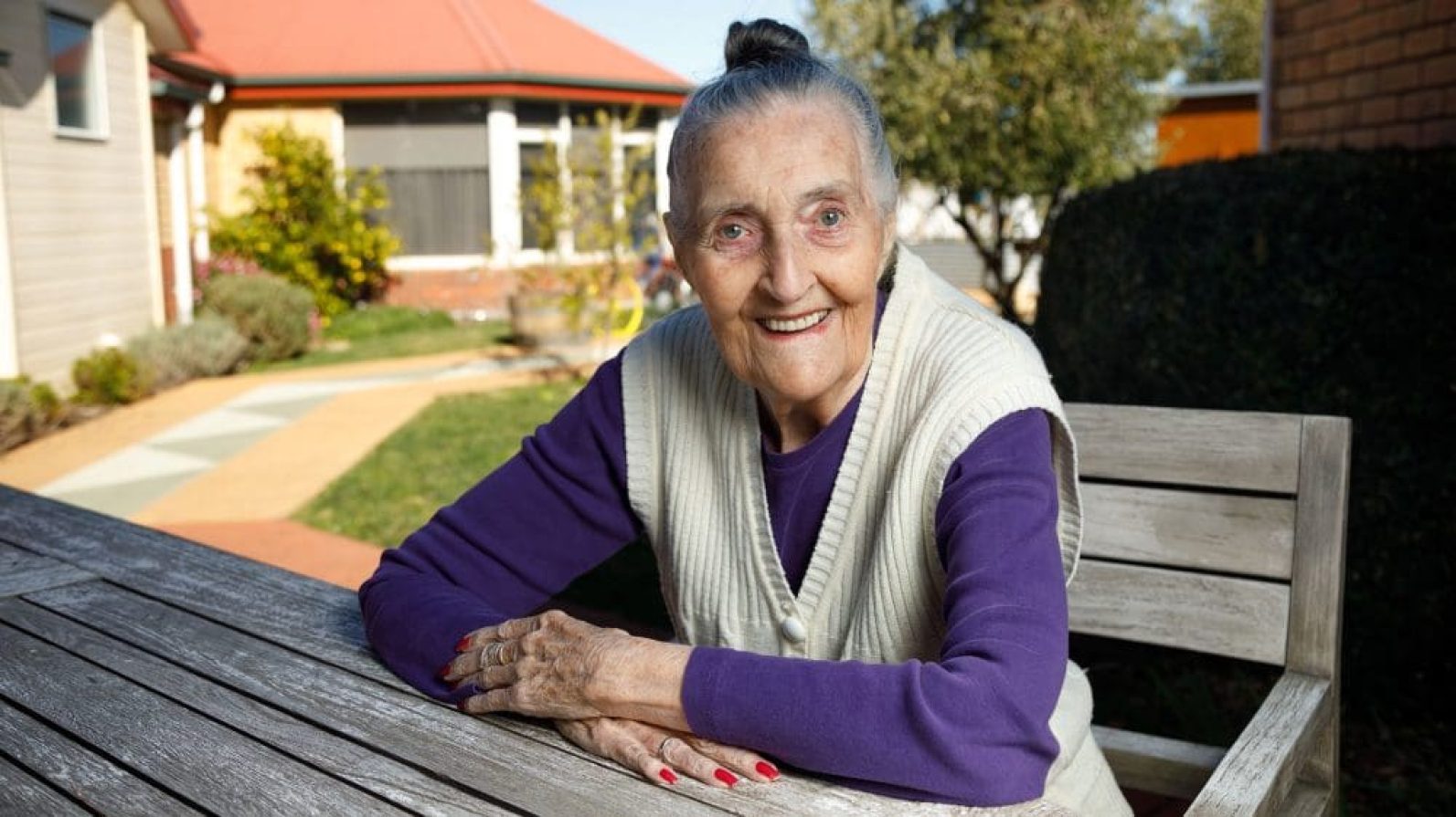 A person sits at a timber outdoor table in a landscaped courtyard, wearing a purple top and cream knitted vest. Residential buildings, pathways, lawns and garden plantings surround the sunny outdoor space, creating a welcoming community environment.