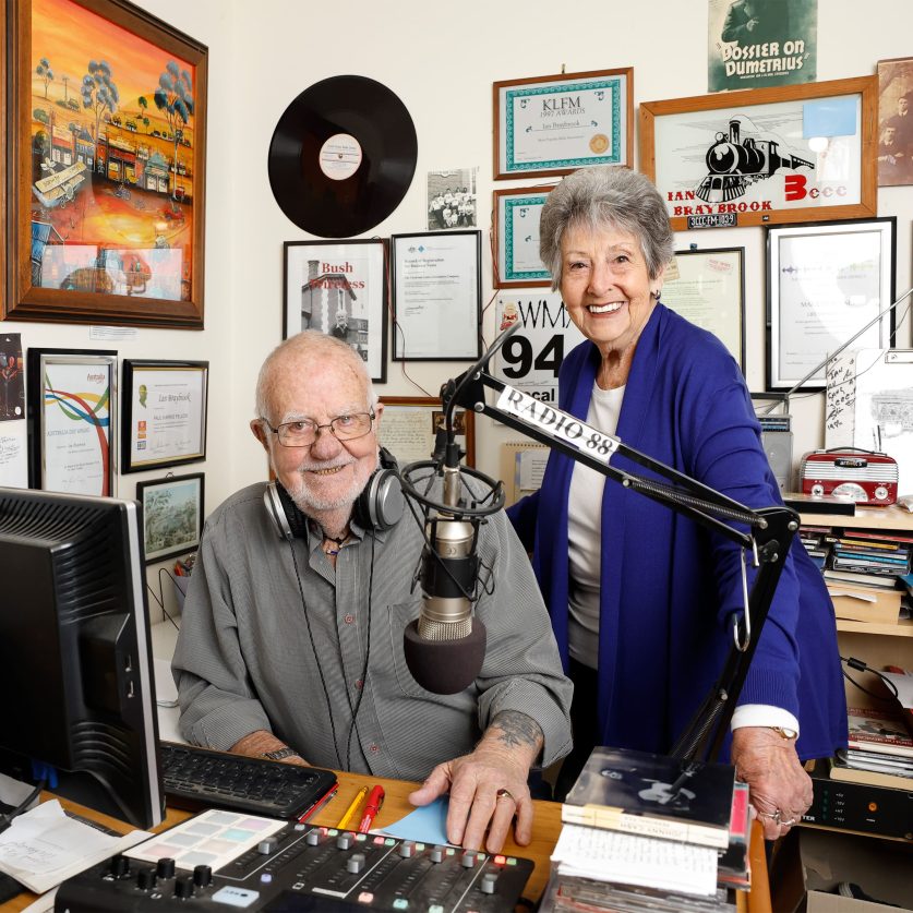 Two older adults in a community radio studio, seated beside a microphone, audio mixing desk and computer. Framed awards, certificates, photographs and radio memorabilia line the walls behind them, highlighting involvement in broadcasting, creative pursuits and community engagement.