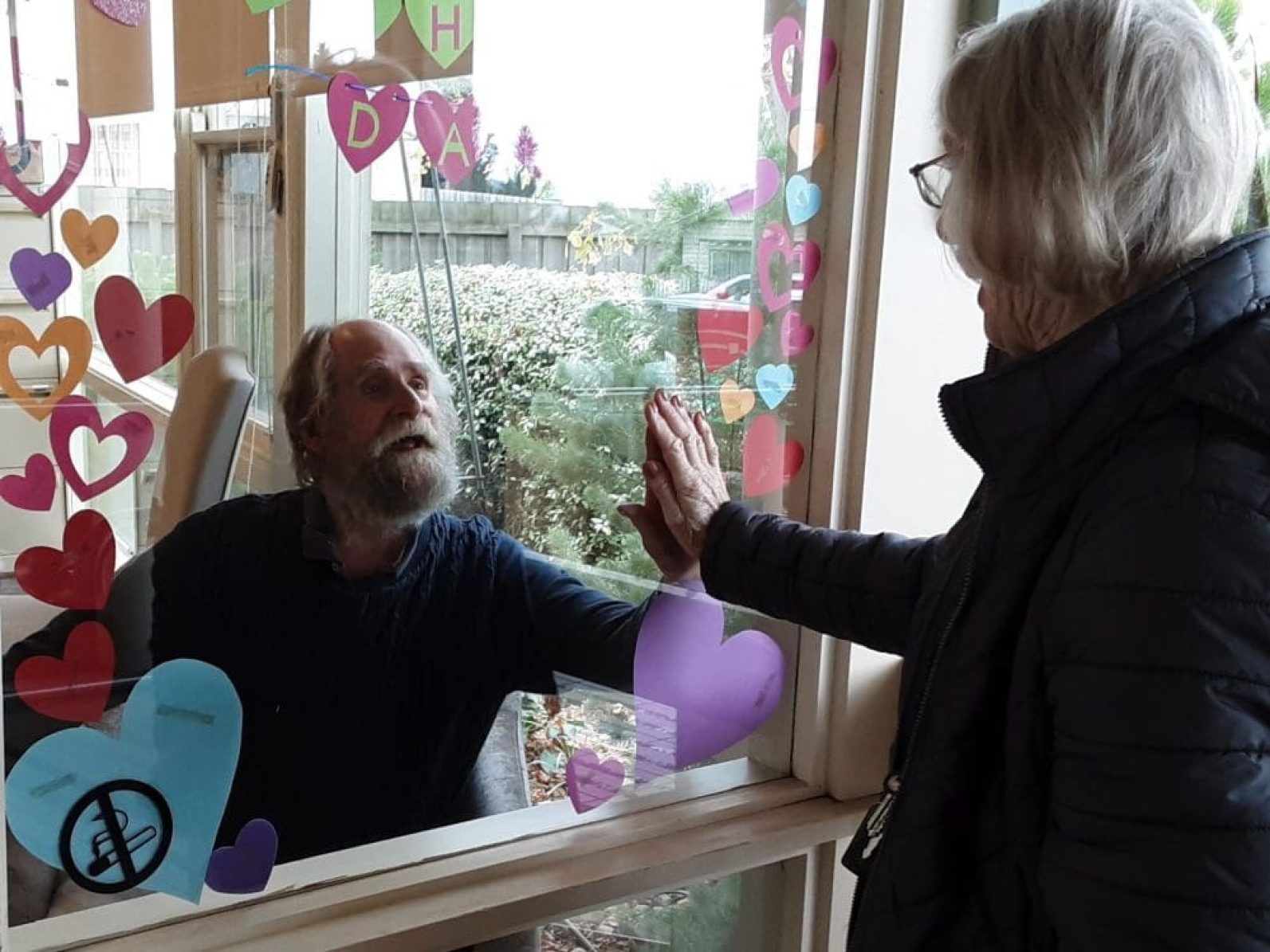 A person stands indoors behind a window decorated with colourful paper hearts, pressing a hand against the glass while greeting another person standing outside. The pair connect through the window, with garden greenery visible beyond the glass, highlighting a moment of social connection and communication during a visit.