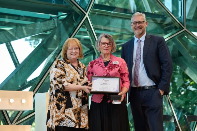 Three people are photographed on stage at the AgeWell Awards. The person in the middle is holding their awards. On the right is Uniting AgeWell CEO Andrew Kinnersly