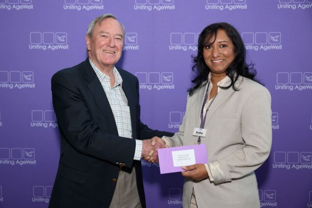 Two people are photographed shaking hands in front of the Uniting AgeWell media wall