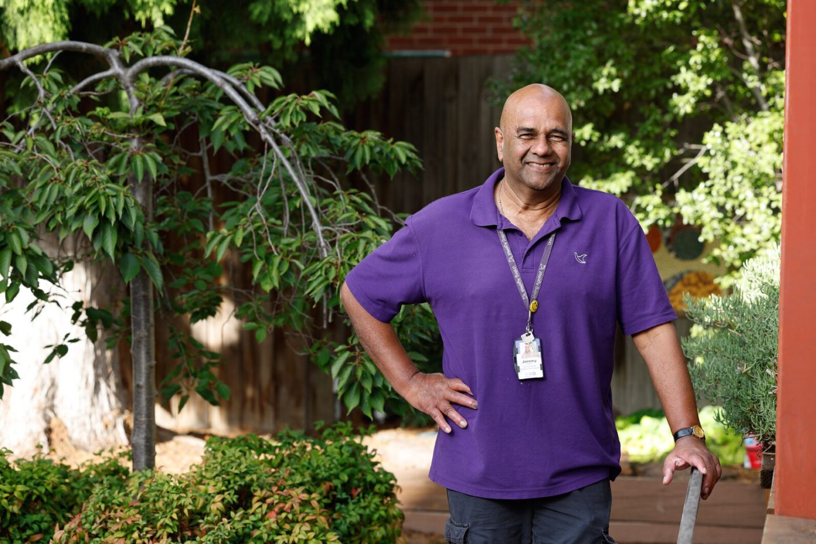 A staff member in a purple Uniting AgeWell polo shirt stands in a leafy garden courtyard, resting one hand on a handrail and the other on their hip. Trees, shrubs and garden beds surround the outdoor space, creating a welcoming and well-maintained environment.