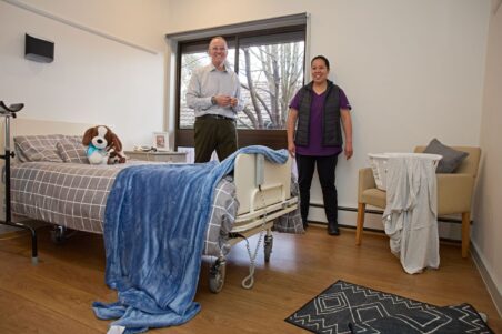A Uniting AgeWell customer and a staff member are standing near the windows in a room. On the bed, there is a plush toy dog also looking at the camera