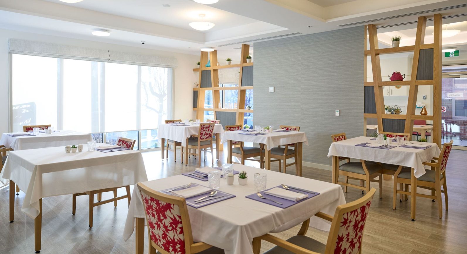 The image captures the dining area at Manor Lakes Community, featuring tables with four chairs at each of them.